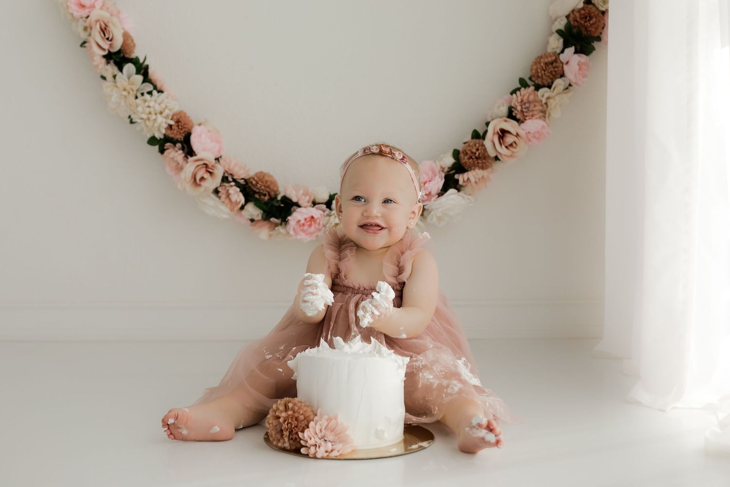 Baby girl in a pink tulle dress near a white window smashing a simple white cake for her studio cake smash portrait session.