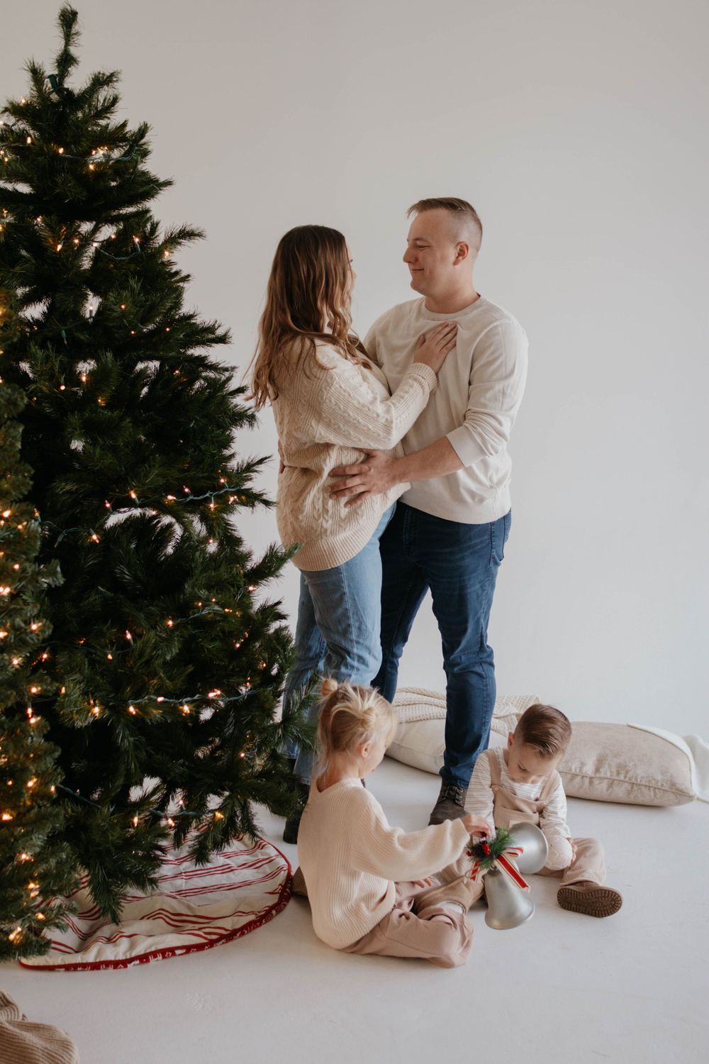 Family decorating Christmas tree together in cozy room with children sitting on floor next to lit evergreen. Studio family photoshoot
