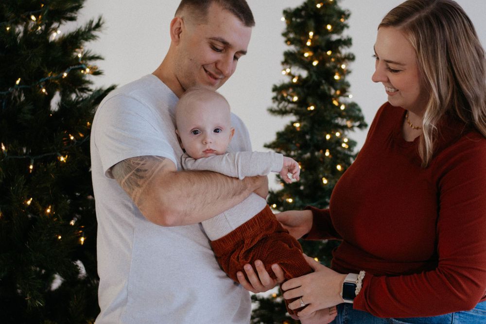 A baby in a white onesie and brown pants snuggles with their parents at holiday family photoshoot in a studio in Colorado