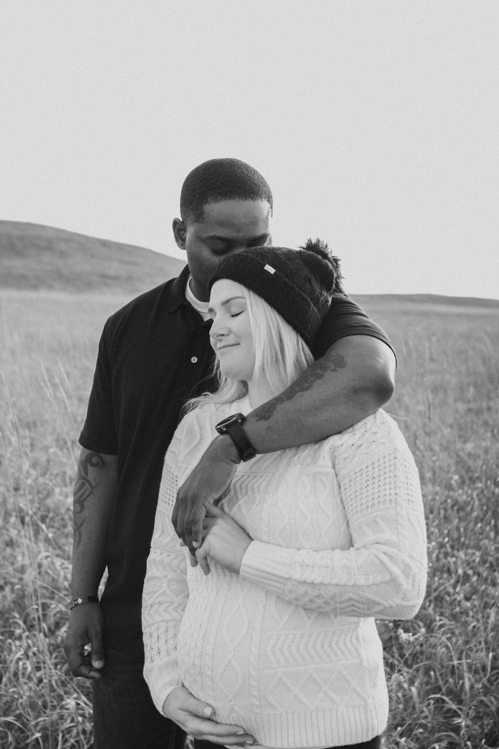A couple shares an intimate moment in a field as they celebrate their pregnancy in a black and white photograph. Maternity family photoshoot
