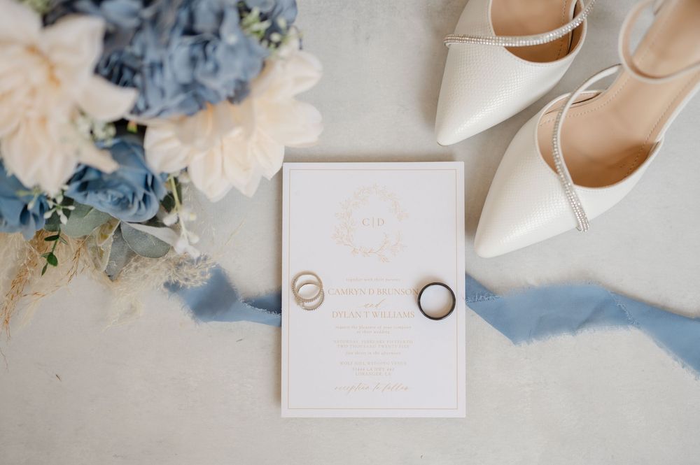 Wedding details flatlay with white invitation card, silver rings, ivory heels and blue and cream floral accents on light surface.
