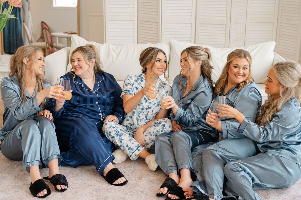Group wearing matching silk pajamas sits together on white couch sharing drinks and laughs during special occasion.