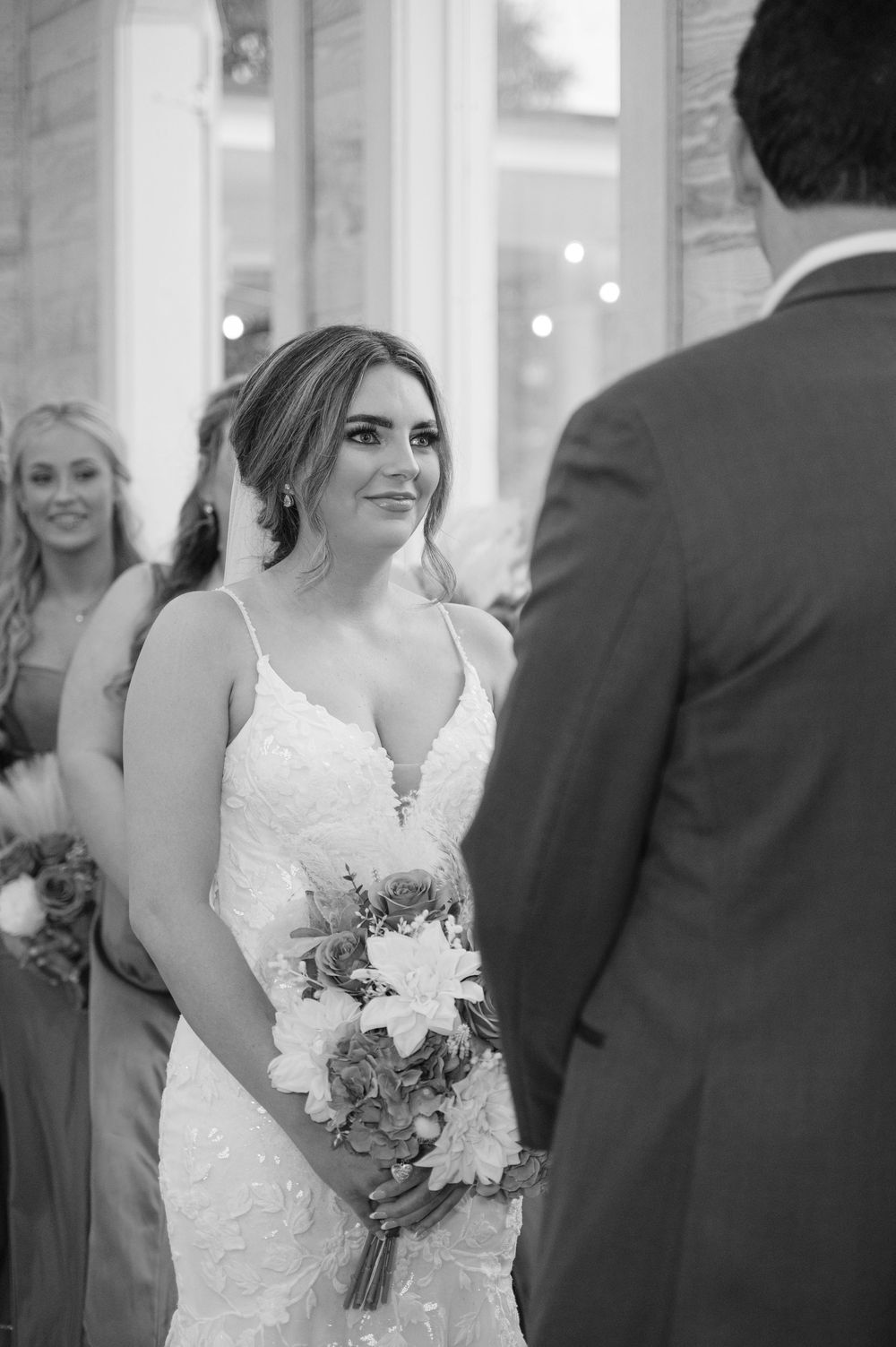 Black and white wedding ceremony photo of a bride in lace dress holding bouquet at the altar.