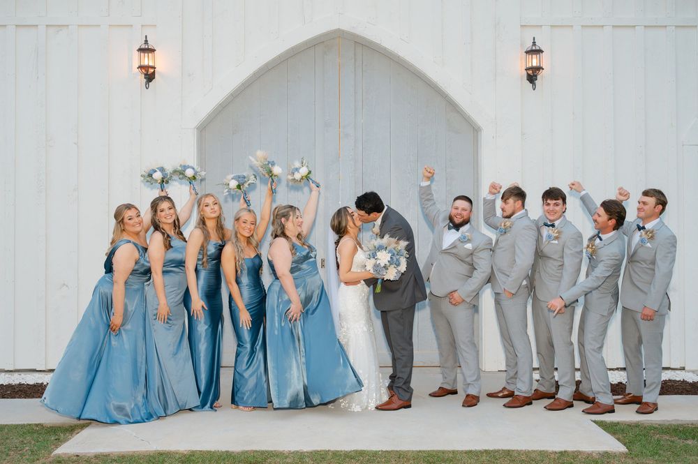 Wedding party poses against white church wall with bridesmaids in blue dresses and groomsmen in gray suits.