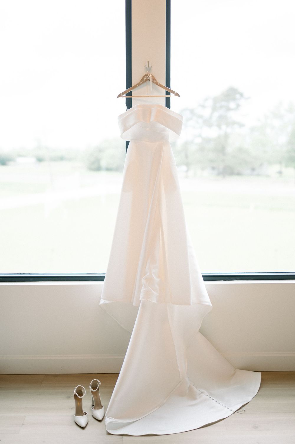 An elegant white wedding dress hangs by a window with matching white heels placed beneath it.