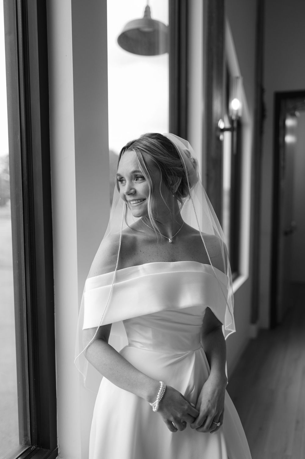 Black and white portrait of a bride in an off-shoulder wedding dress and veil standing by a window in natural light.