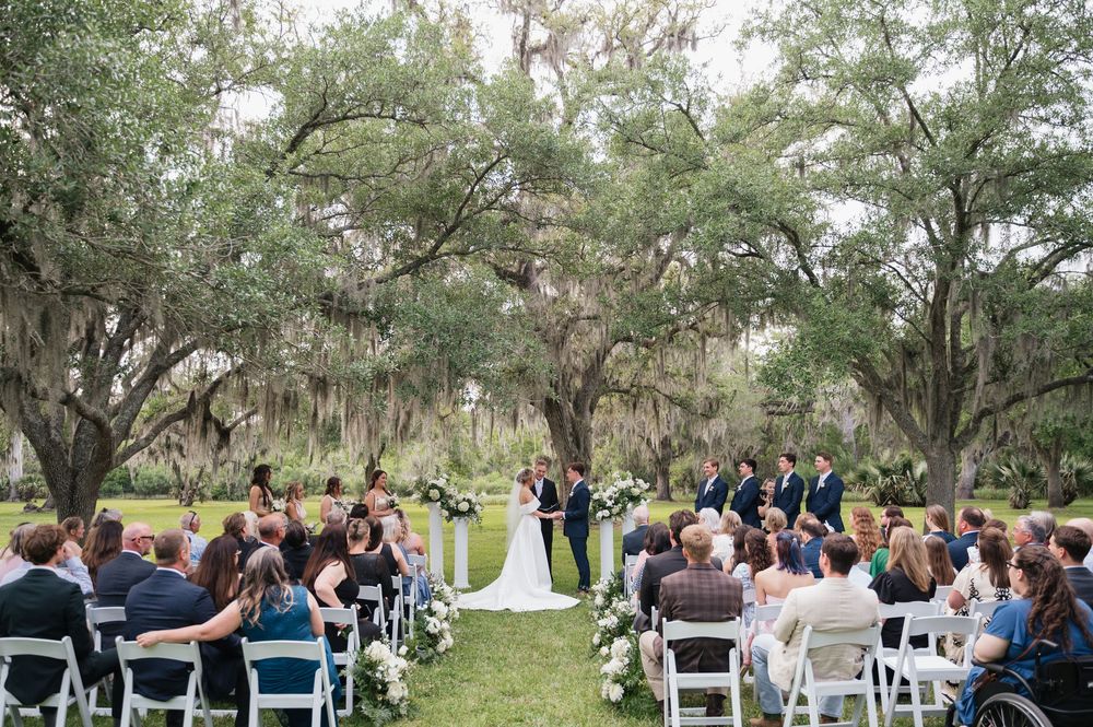 Outdoor wedding ceremony under old oak trees draped with Spanish moss as guests watch from white folding chairs.
