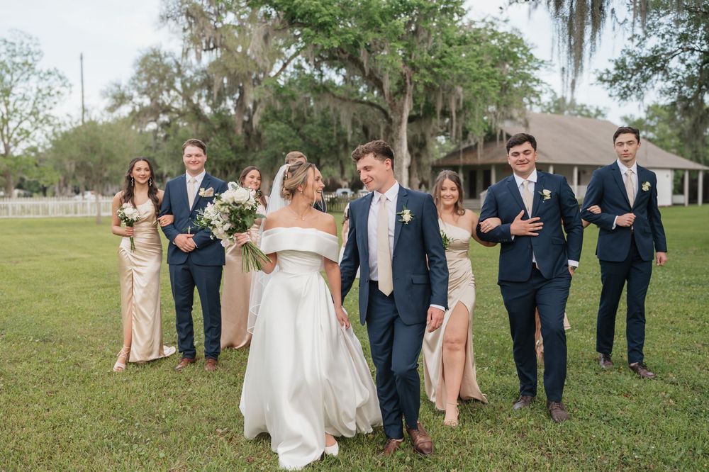 Wedding party walks on green lawn with bridesmaids in gold dresses and groomsmen in navy suits at outdoor venue.