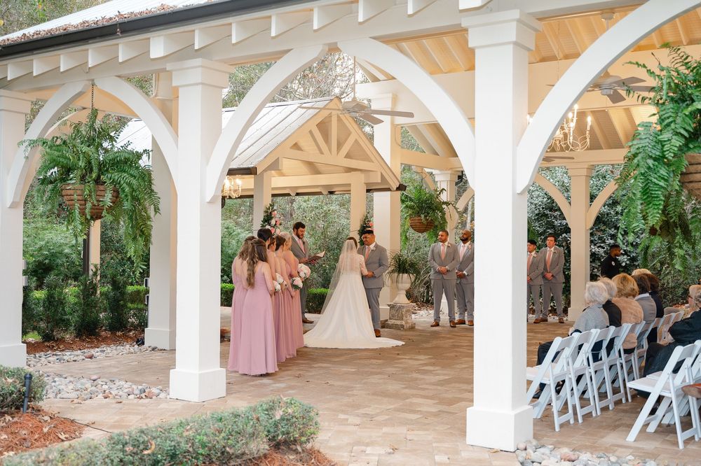 An outdoor wedding ceremony takes place under elegant white arches adorned with greenery and flowers.