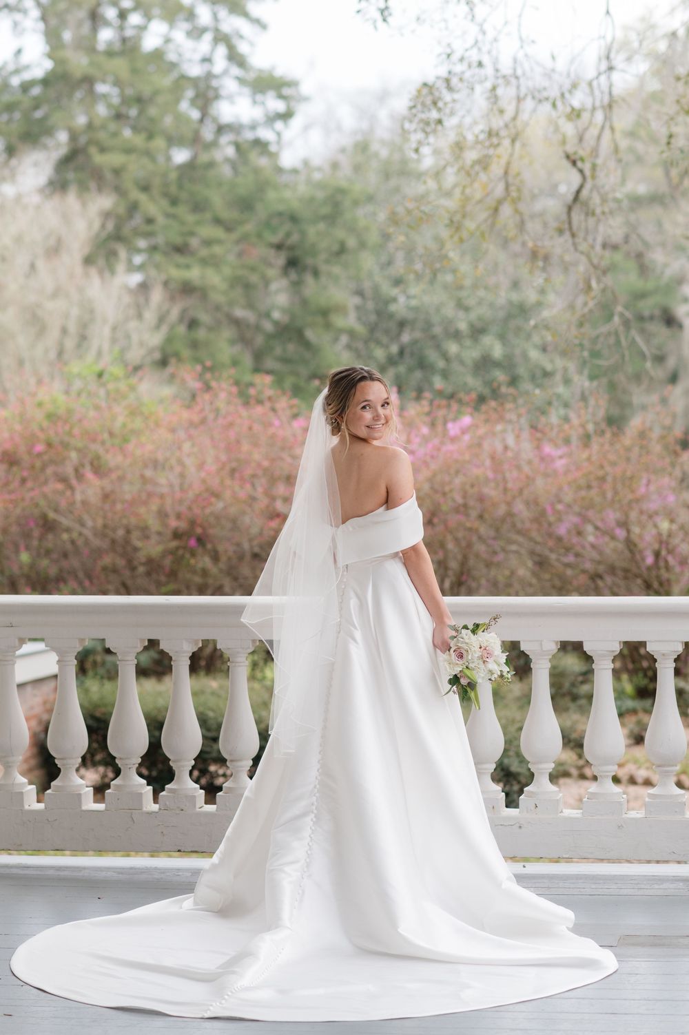 A bride in an elegant off-shoulder white wedding gown with long train stands on a balcony surrounded by autumn foliage.