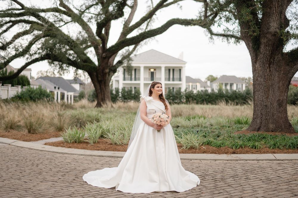 Elegant bride in flowing white gown holds bouquet while standing on brick path under majestic oak trees.