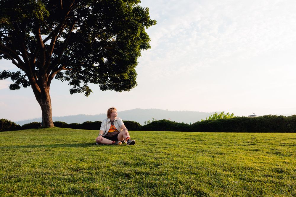 A woman sits on the grass in front of a tree, enjoying a serene moment during her senior session.