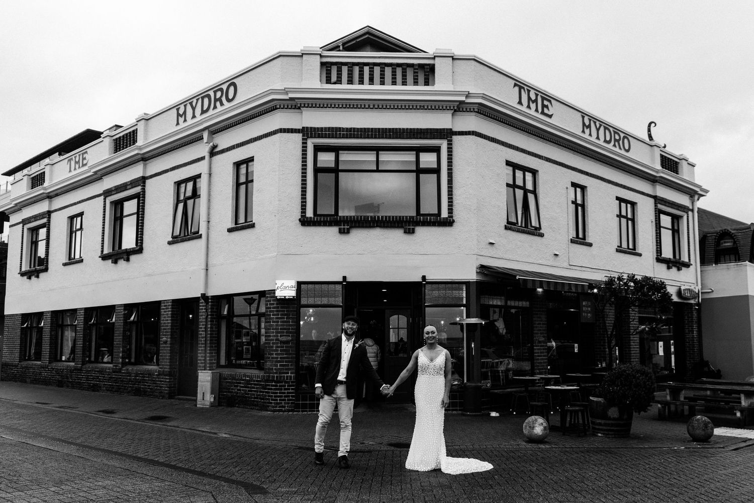 A black and white wedding photo of a couple outside a vintage corner building.