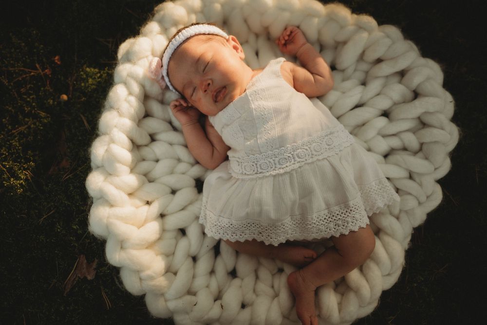 Sleeping baby in white lace dress on chunky knit cream wool blanket.