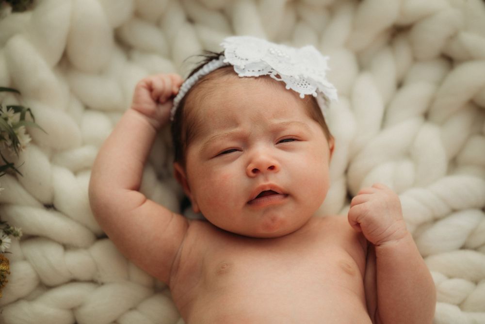 Newborn baby lying on chunky knit cream blanket wearing white lace headband.