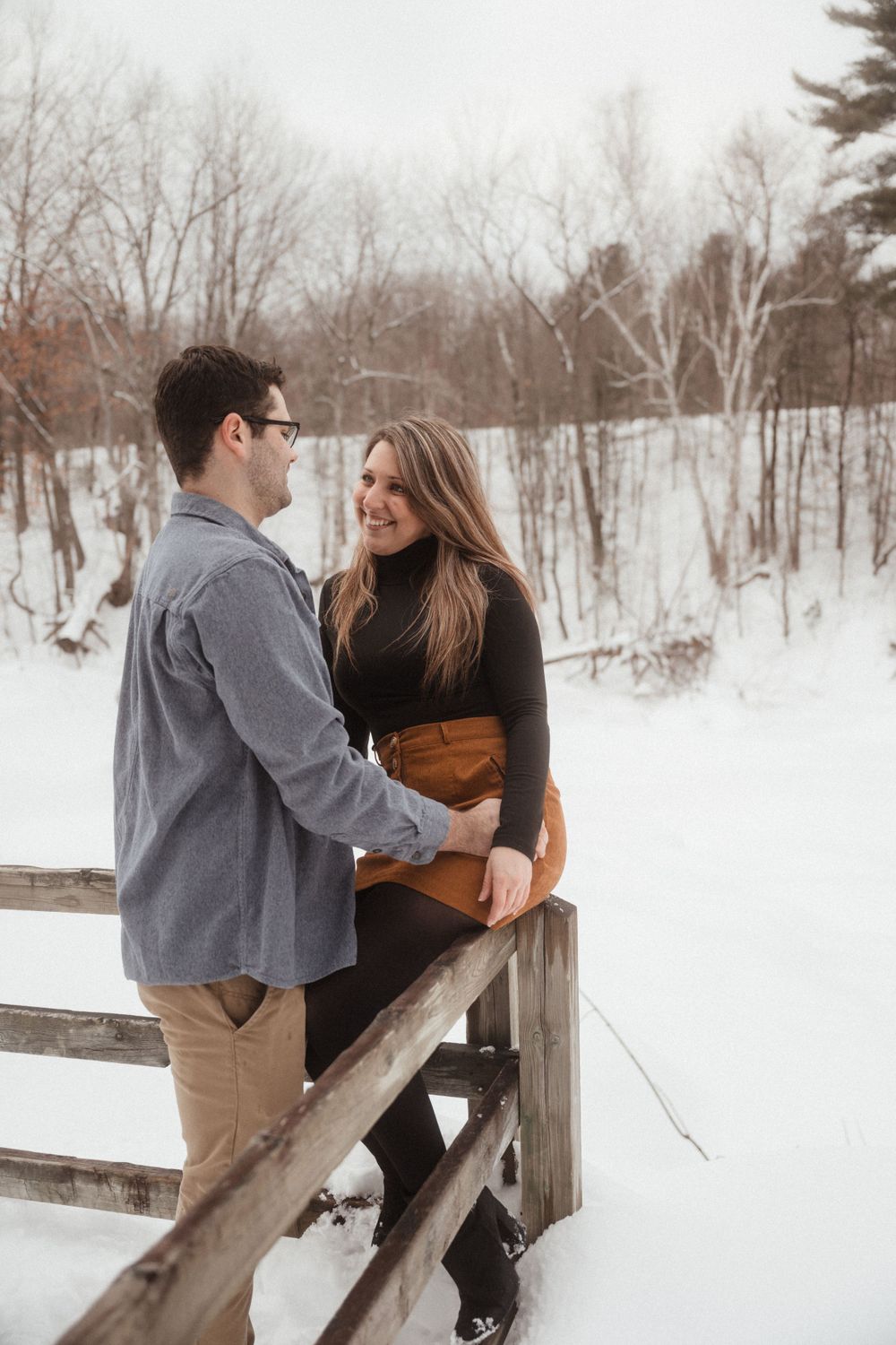 Couple shares an intimate moment on a snowy wooden fence with a rustic bridge in the distance.