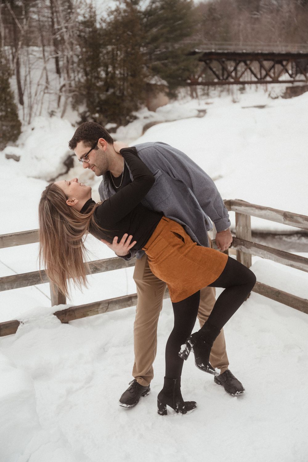 A couple performs a romantic dip pose on a snow-covered wooden bridge during winter.