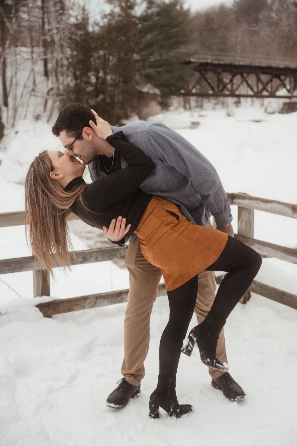 A romantic winter photo of a couple sharing a dip kiss on a wooden bridge with snow and trees in the background.