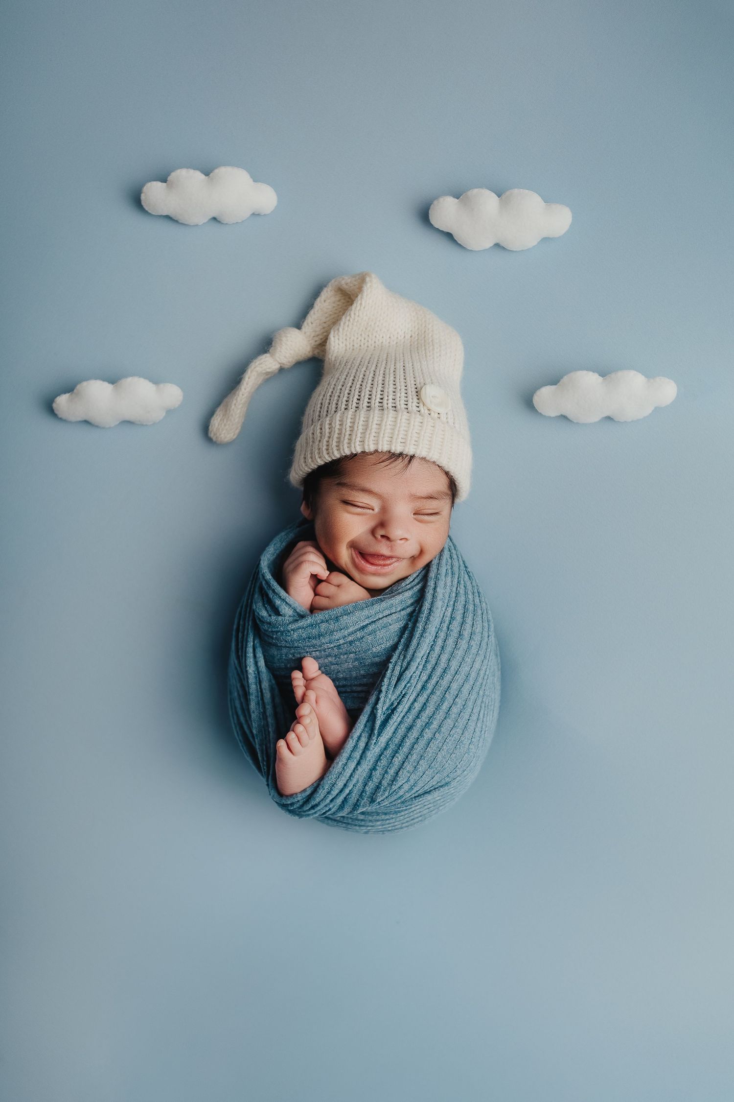 Newborn baby wrapped in blue swaddle wearing white knit hat smiles while sleeping against blue backdrop with cloud decorations.