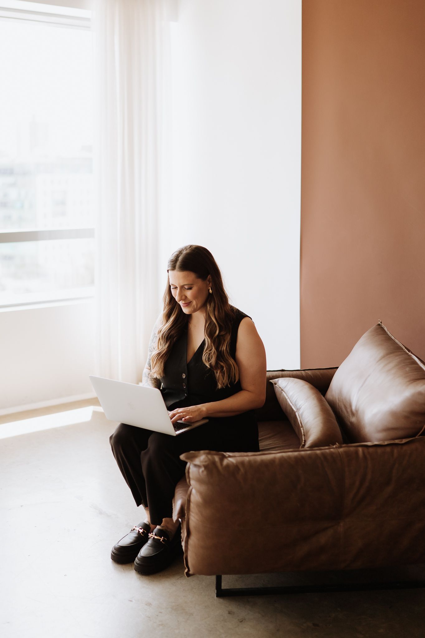 Woman working on laptop while relaxing on brown leather couch near window.