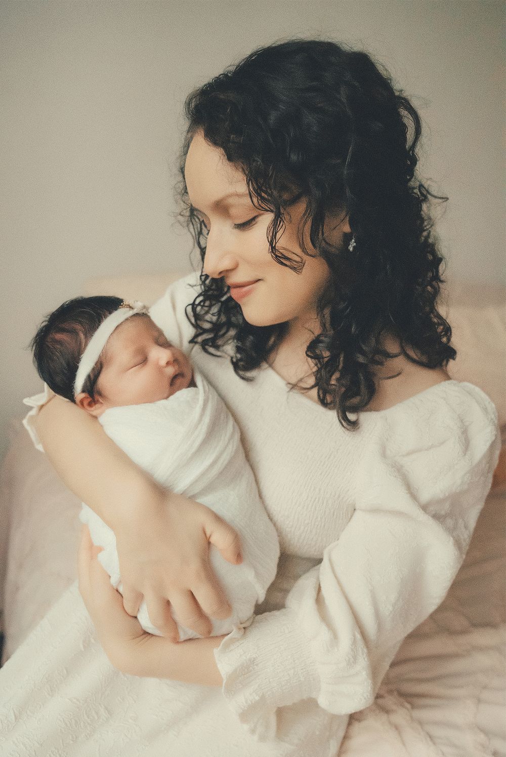 A woman in a white dress tenderly holds and gazes at her sleeping newborn baby swaddled in white cloth.