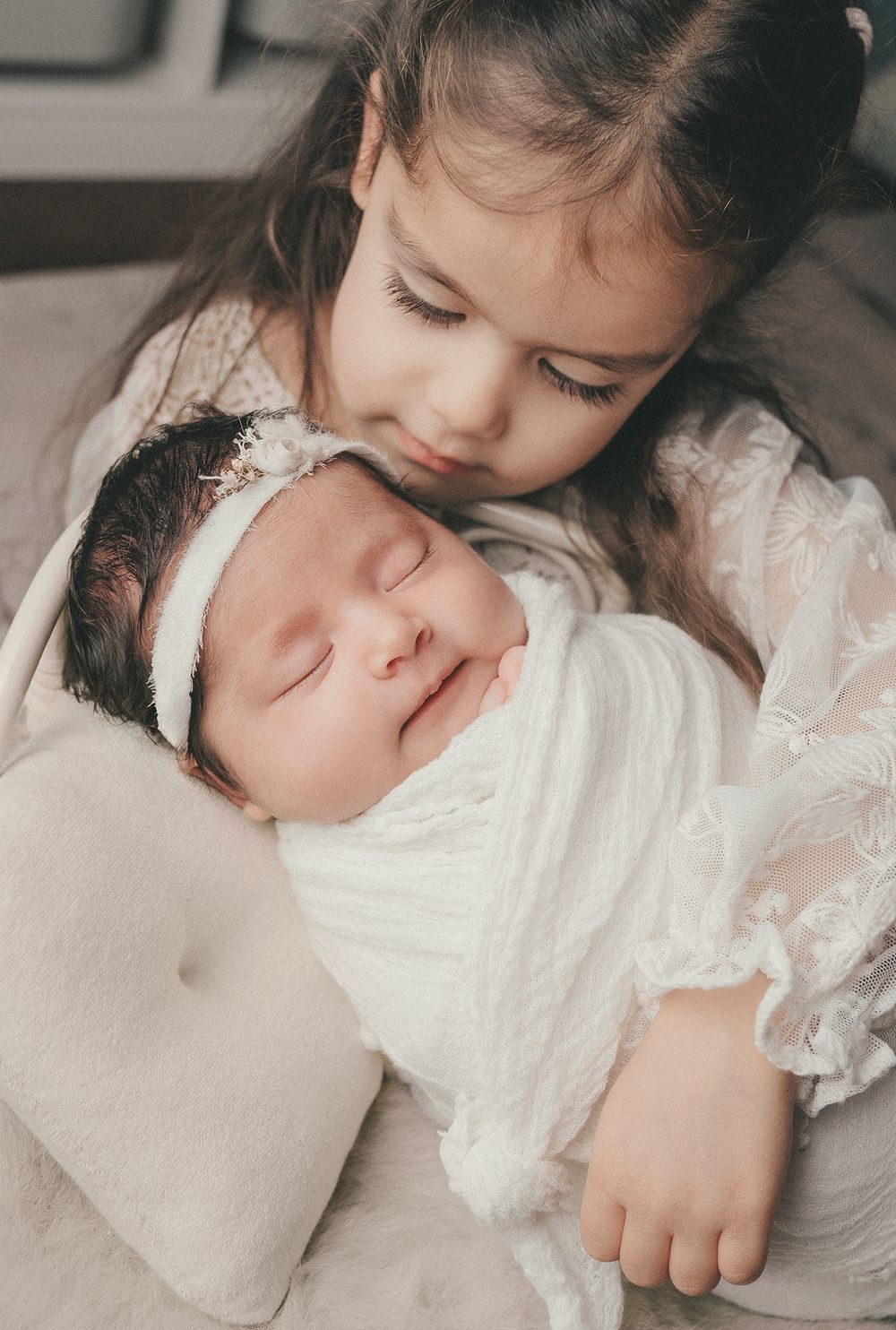 A young child in lace dress cuddles with a sleeping newborn baby swaddled in white.