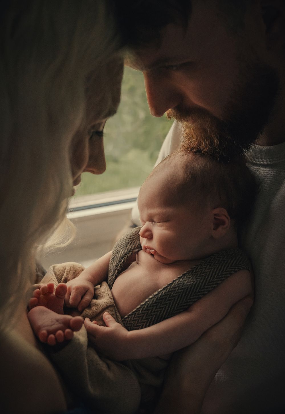Intimate family moment as parents lovingly gaze at their sleeping newborn baby in soft natural light.