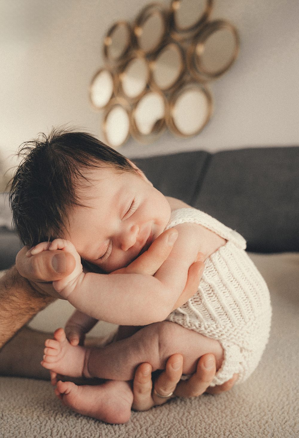 A sleeping newborn baby wearing a white knit romper rests curled up on a gray couch beneath circular wall decor.