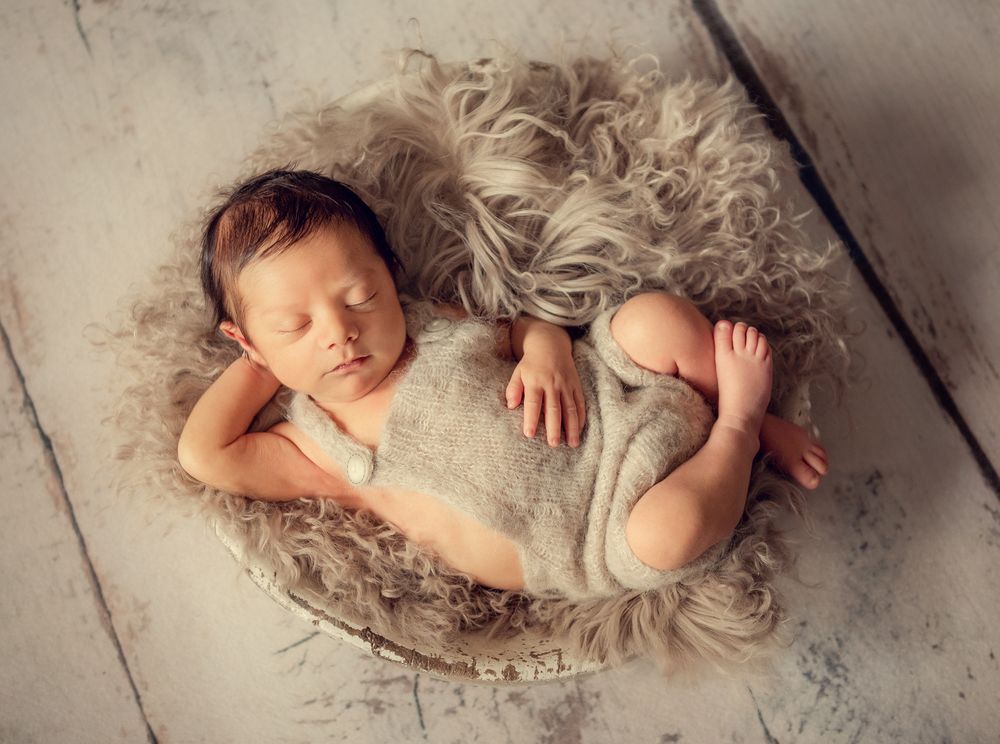 newborn photo session of the baby wearing knitted romper posing in a wooden bowl stuffed with a fur photographed at home