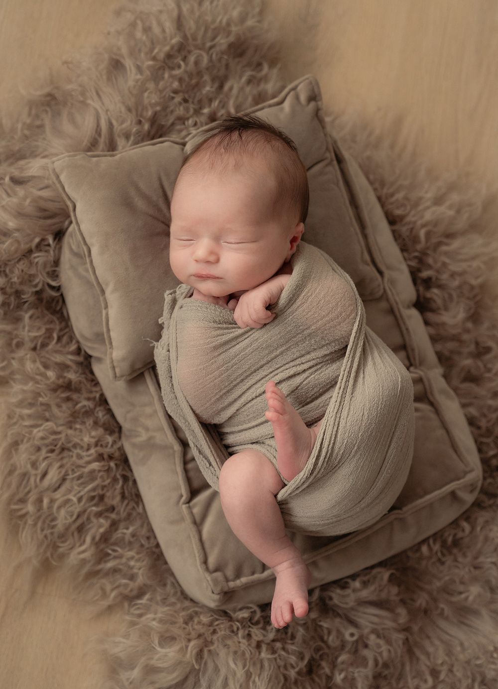 Sleeping newborn wrapped in soft beige fabric lying on textured gray pillow surrounded by fluffy sheepskin.