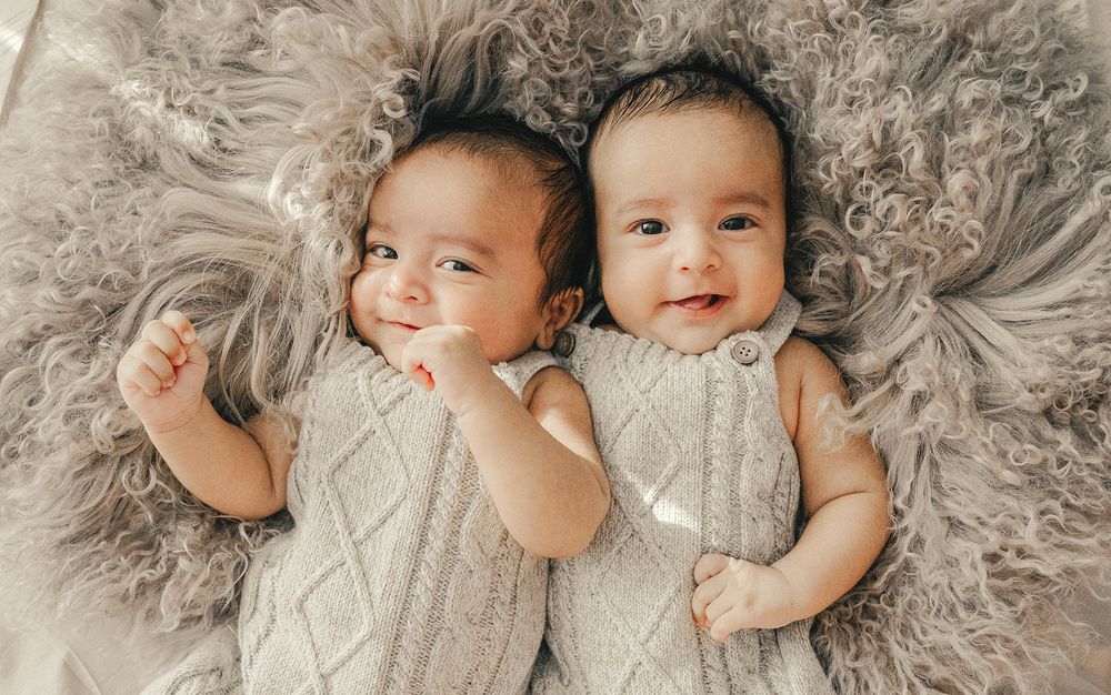 Two adorable babies lying side by side on a soft gray fur blanket wearing matching knit cream outfits.