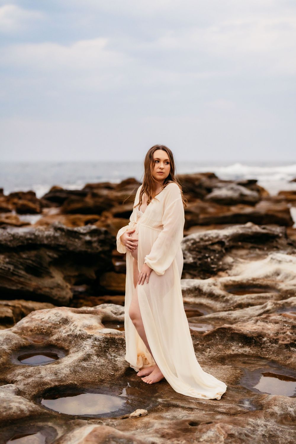 Woman in flowing white dress poses on rocky coastal outcrop during dramatic oceanside maternity photoshoot.