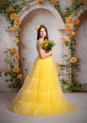 A model in a flowing yellow tulle gown and floral crown poses against a romantic archway adorned with peach roses.