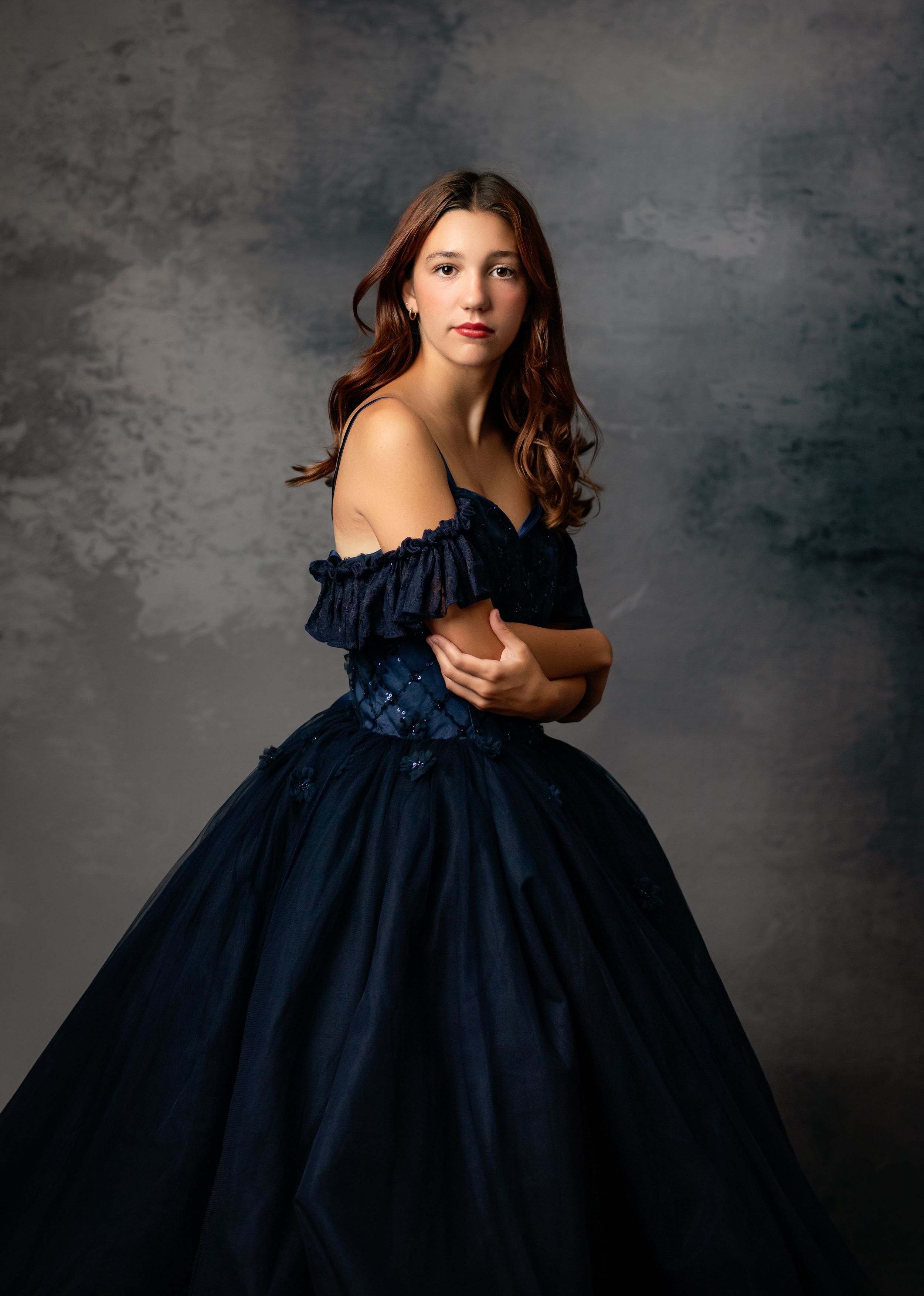 A woman in an elegant navy blue ball gown poses dramatically against a dark grey textured backdrop.