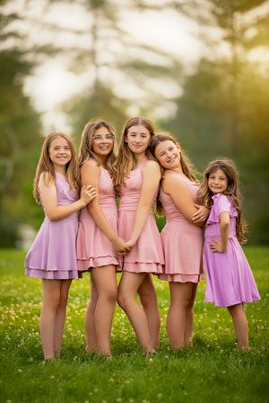 A group of five people wearing coordinating pink dresses stand together on grass in warm sunlight.