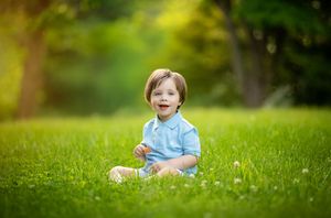 Young child in blue shirt sitting and laughing on green grass in a sunlit outdoor setting during spring or summer.