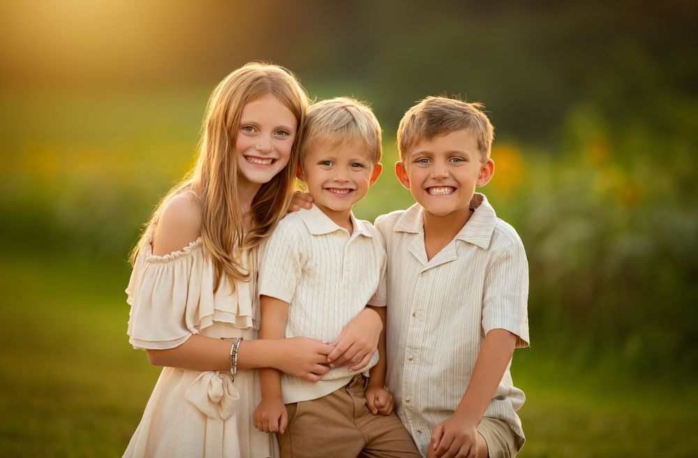 Three children wearing light-colored outfits sitting together outdoors during golden hour in a sunny field.