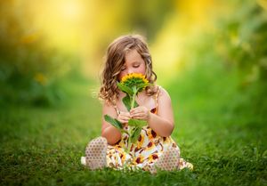 A child in polka dot boots sits in lush grass smelling bright yellow sunflowers on a sunny summer day.