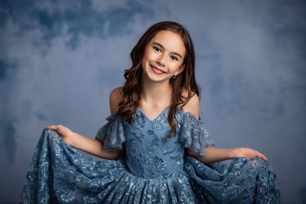 Young person wearing an elegant blue lace dress poses against a textured blue background with a bright smile.