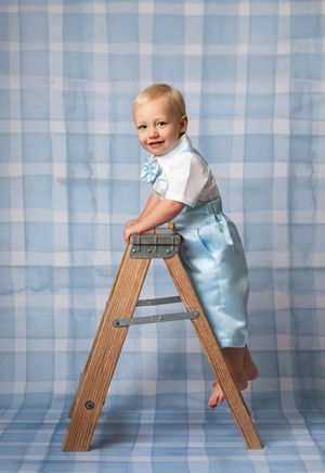 A small child in light blue and white outfit climbs on a wooden stepladder against a blue plaid backdrop.