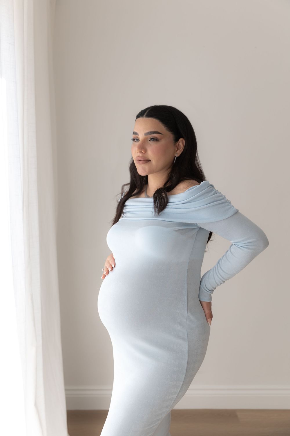 Pregnant woman in light blue long sleeve maternity dress poses by window in soft natural light.