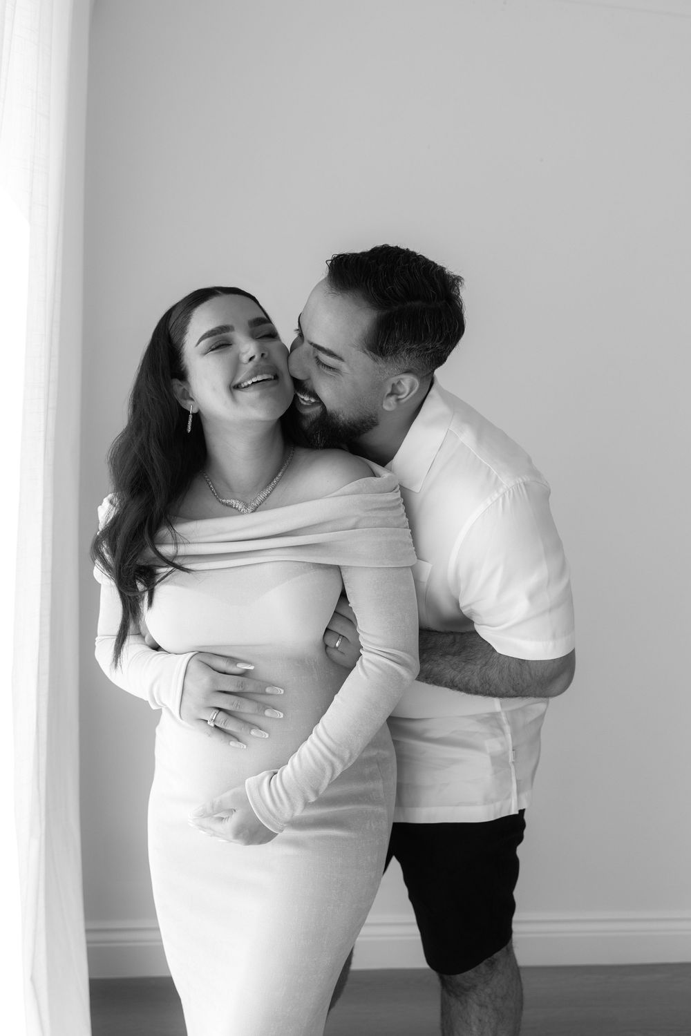 Intimate black and white maternity photo of couple embracing and laughing by window.