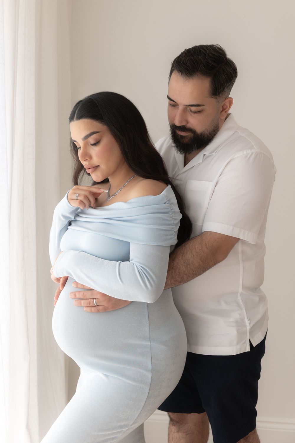Tender moment between expecting couple as they pose together by window in matching light colored outfits.