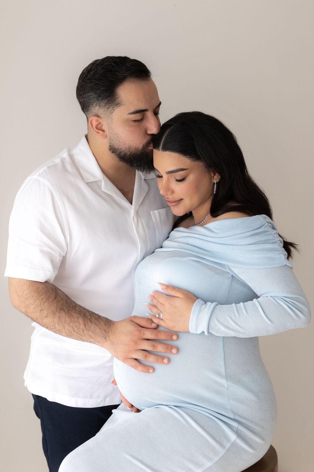 A romantic couple embraces tenderly while wearing white and blue clothing against a neutral background.
