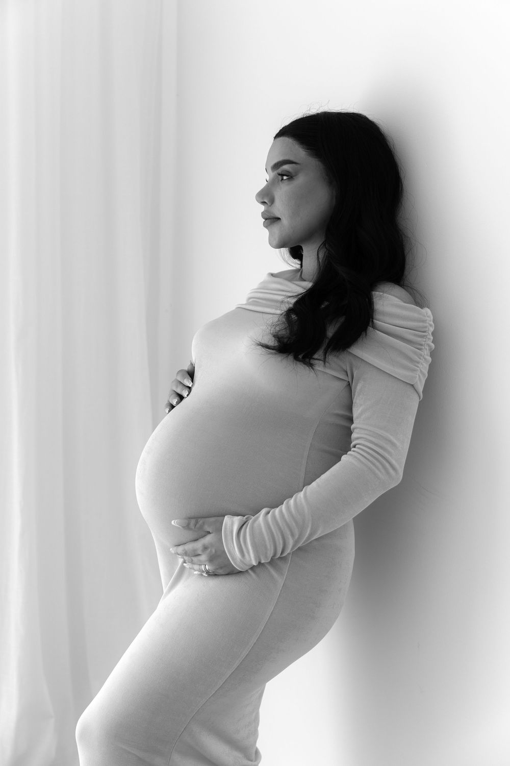 Black and white maternity photo of a woman in a fitted white dress poses against a light backdrop.