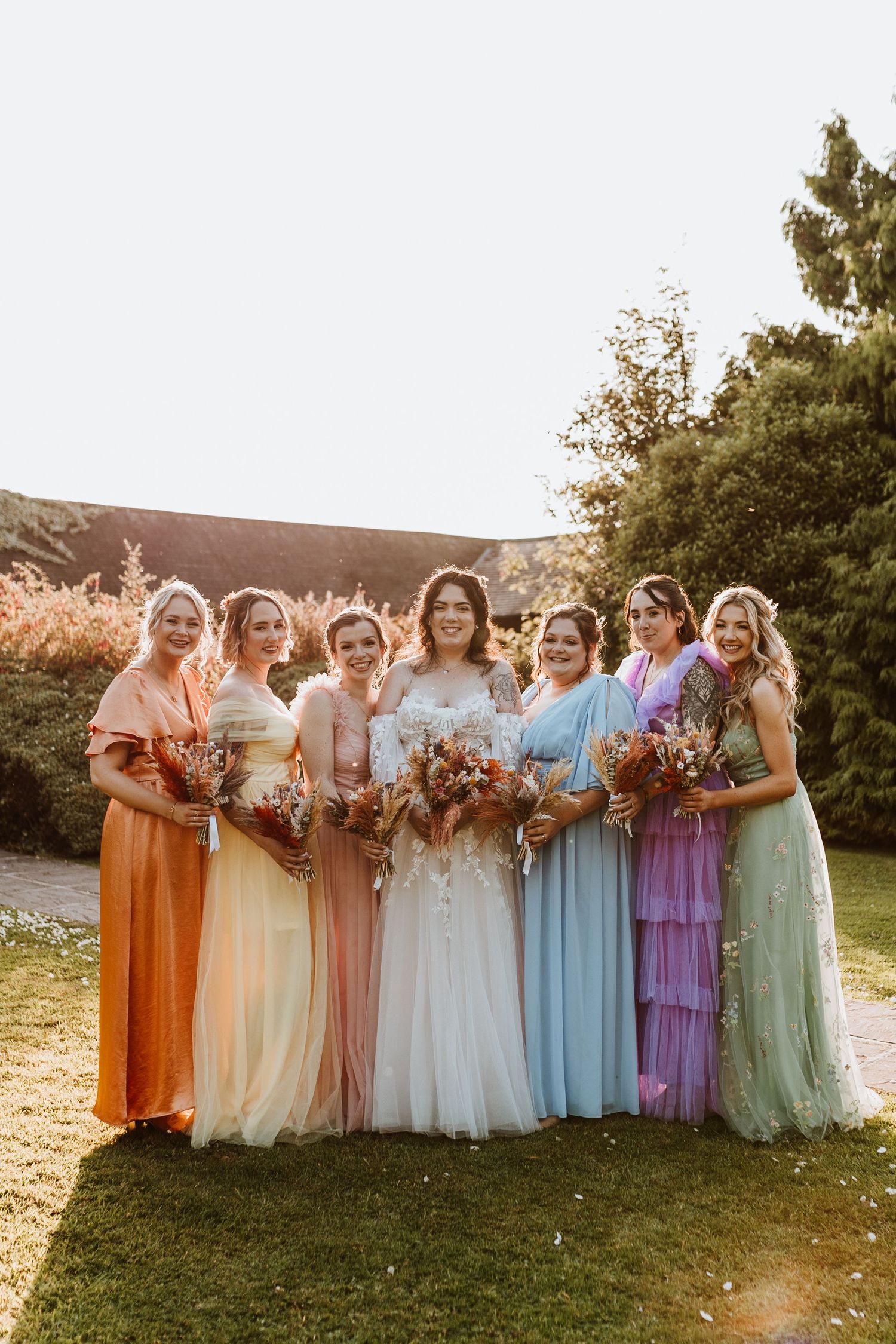 Bridesmaids in rainbow pastel dresses and the bride pose together at sunset in a garden setting.