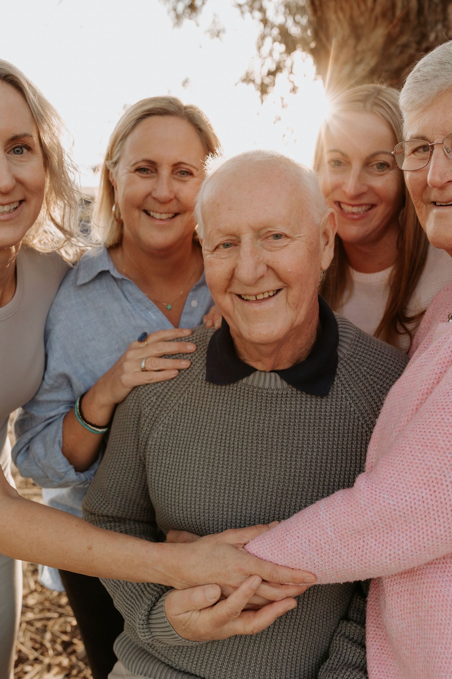 A group of people gather close together outdoors, smiling and embracing in warm sunlight.