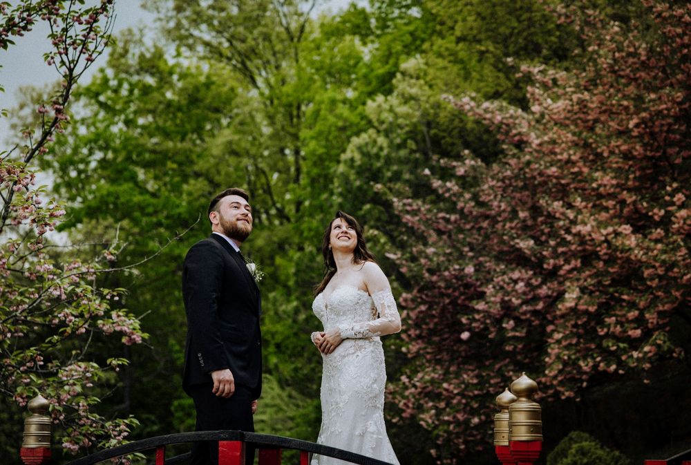A couple stands together amid blooming trees in an outdoor garden setting with decorative lanterns nearby.