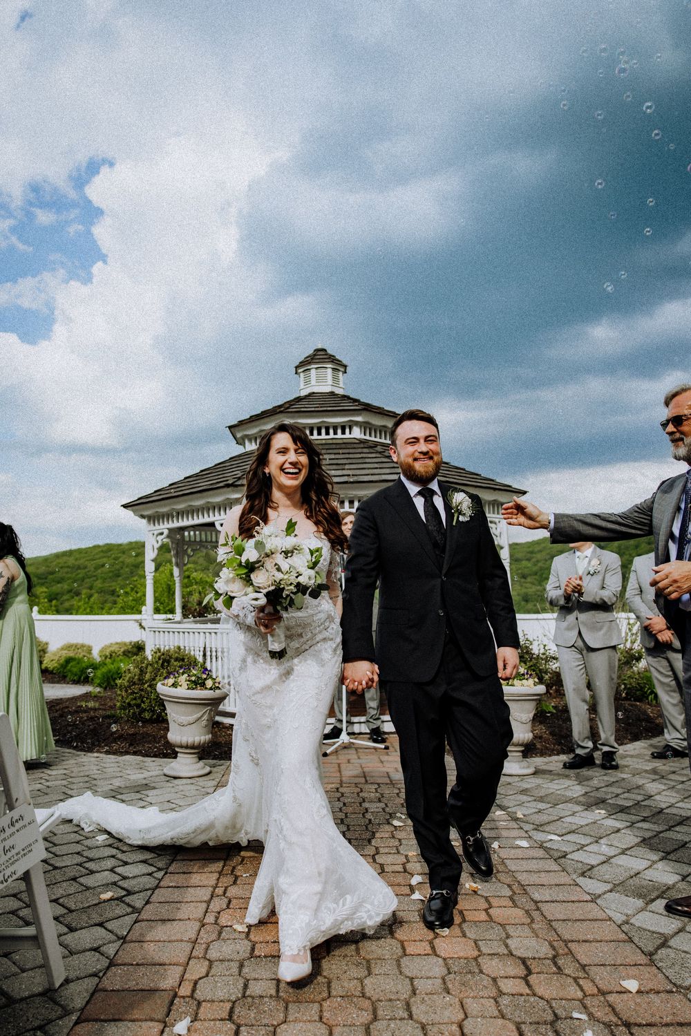 Happy couple walks together on stone path after outdoor wedding ceremony with white gazebo and cloudy sky in background.