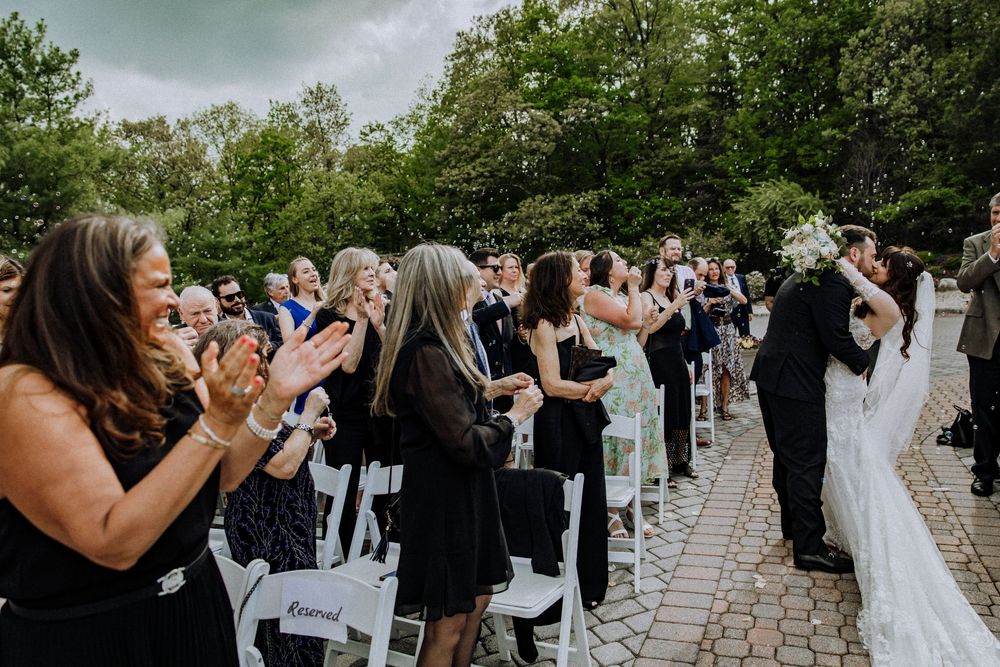 Wedding guests in black attire stand and applaud as newlyweds celebrate during an outdoor ceremony on a brick patio.